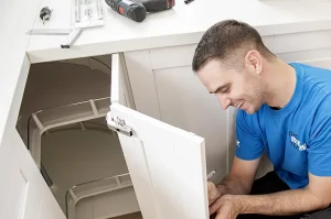 Hilfe beim Küchenaufbau: Ein junger Monteur in blauem T-Shirt kniet vor einem geöffneten Küchenschrank und hält einen Stift in der Hand.